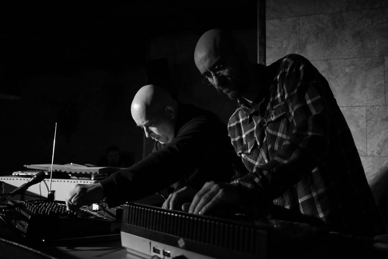 Fotografia czarno-biała. Kadr na dwóch mężczyzn pracujących przy sprzęcie muzycznym.