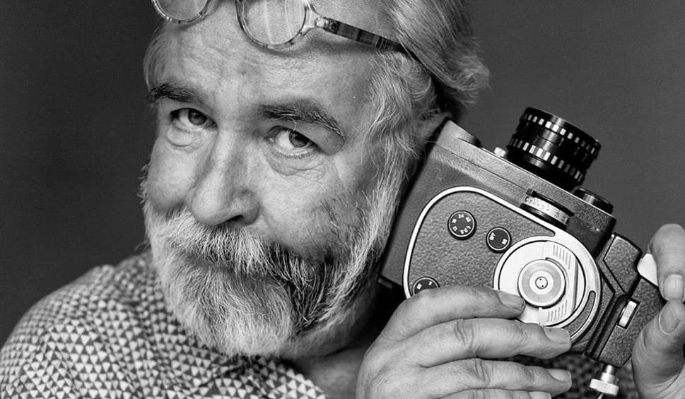 Fotografia czarno-biała. Kadr na twarz Yacha Paszkiewicza przytulający aparat analogowy.