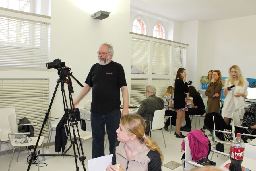Fotografia kolorowa. Wnętrze warszatatowe. Kadr na uczestników warsztatów oraz prowadzącego Yacha Paszkiewicza wraz ze sprzętem nagraniowym.