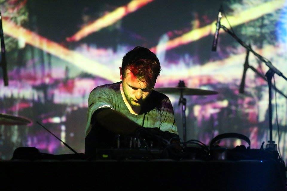 Fotografia kolorowa. Kadr na mężczyznę stojącego nad sprzętem do muzyki elektronicznej. W tle wyświetlane wizualizacje