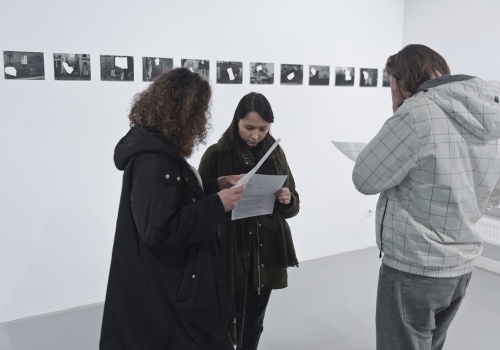 Zdjęcie, trójka uczestników wernisażu przegląda notatki związane z wystawą. W tle znajdują się zdjęcia Hélène Thiennot prezentowane na ścianie Sali wystawienniczej.