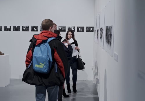 Zdjęcie, kilkoro uczestników wernisażu ogląda prace artystki prezentowane na ścianie Sali wystawienniczej.
