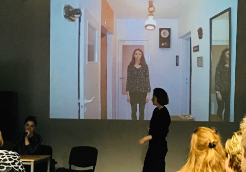 Zdjęcie, wnętrze Parakina, w trakcie trwania wykładu. Na ścianie wyświetlana jest prezentacja. Po lewej i prawej stronie kadru siedzą uczestnicy, obróceni tyłem do obiektywu. Środkiem Sali przechodzi kobieta, która znajduje się w centrum kadru.