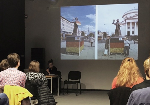 Zdjęcie, wnętrze Parakina, w trakcie trwania wykładu. Na ścianie wyświetlana jest prezentacja. Po lewej i prawej stronie kadru siedzą uczestnicy, obróceni tyłem do obiektywu.
