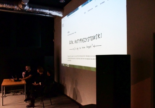 Zdjęcie, wnętrze Parakina w trakcie trwania wykładu. W lewym rogu kadru znajduje się stół przy którym siedzą trzy osoby. Na ścianie wyświetlana jest prezentacja – czarne napisy na białym tle. 