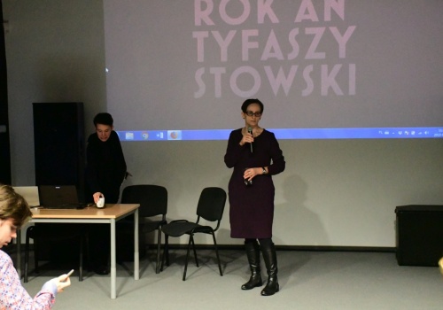 Zdjęcie, w centrum kadru znajduje się Dyrektor CSW ŁAŹNIA Jadwiga Charzyńska, uwieczniona w trakcie wypowiadania się. Po lewej stronie stoi stolik za którym znajduje się jeden z uczestników wykładu. Na ścianie wyświetlana jest prezentacja. 