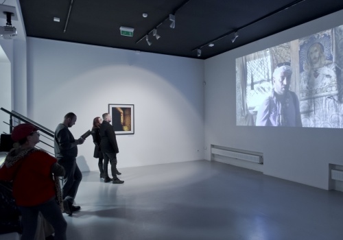 Zdjęcie, wnętrze  Sali Wystawienniczej. Na ścianie po lewej stronie prezentowana jest fotografia Jerzego Wierzbickiego, a po prawej stronie wyświetlany jest film artysty. Film ogląda kilkoro uczestników wernisażu.