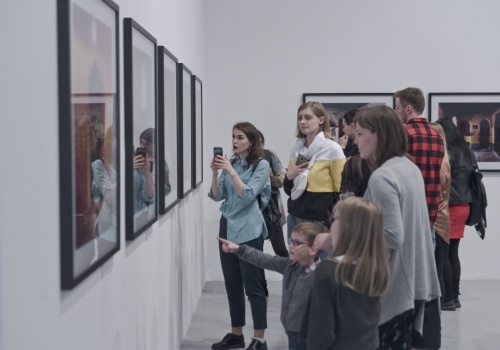 Zdjęcie, licznie zebrani uczestnicy oglądają prezentowane prace artysty we wnętrzu Sali Wystawienniczej. Jedna z uczestniczek podchodzi bliżej i robi zdjęcie telefonem.