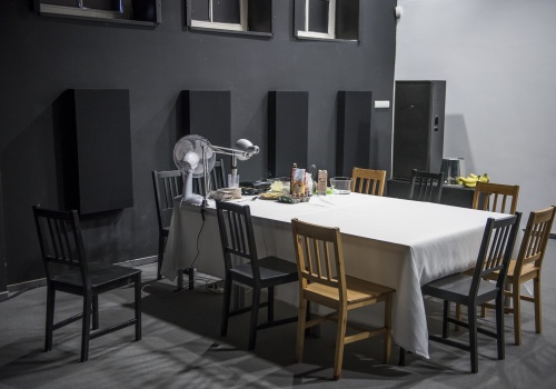 Zdjęcie, widok na stół, który jest elementem performansu Honoraty Martin w Parakinie CSW ŁAŹNIA 1 na Dolnym Mieście. Stół przykryty jest białym obrusem z kilkoma produktami spożywczymi na nim. Przy stole znajdują się krzesła.