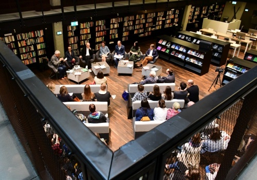 Zdjęcie, widok z góry na osoby prowadzące dyskusje. Przed nimi znajdują się zebrani słuchacze. Za nimi widnieje długi, ciemny regał z książkami. W kadrze mieści się cała przestrzeń biblioteki.