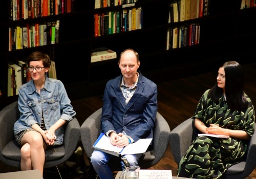 Zdjęcie, zbliżenie na trójkę osób prowadzących dyskusję. Za nimi znajduje się regał z książkami.