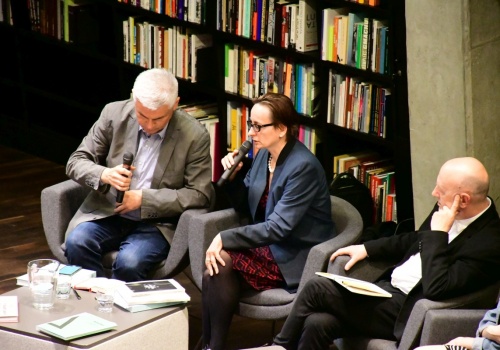 Zdjęcie, widok od boku na osoby prowadzące dyskusje. W centrum kadru znajduje się Dyrektor CSW ŁAŹNIA – Jadwiga Charzyńska, która wypowiada się do mikrofonu.
