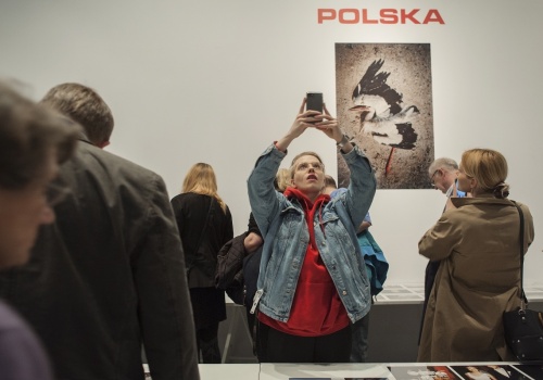 Zdjęcie, licznie zebrani uczestnicy wernisażu oglądają prace Michała Szlagi. Jedna z uczestniczek robi zdjęcia telefonem. W tle, na ścianie, znajduje się fotografia martwego bociana, a nad nią czerwony napis POLSKA.