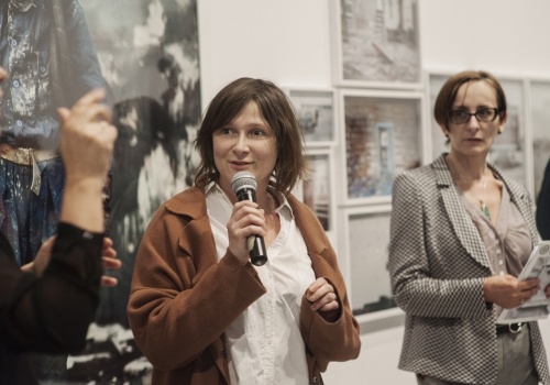 Zdjęcie, zbliżenie na wypowiadającą się do mikrofonu kuratorkę wystawy – Annę Mituś. Towarzyszy jej Dyrektor CSW ŁAŹNIA – Jadwiga Charzyńska.