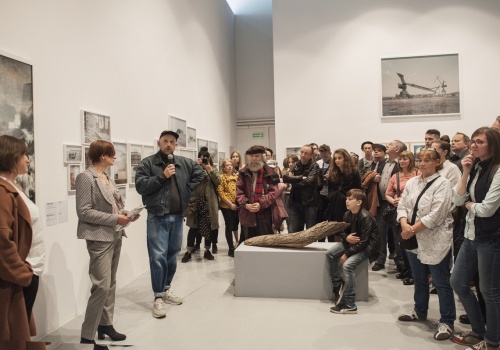 Zdjęcie, po lewej stronie kadru znajduje się kuratorka wystawy, Dyrektor CSW ŁAŹNIA oraz artysta – Michał Szlaga, wypowiadający się do mikrofonu. Po prawej stronie znajdują się licznie zebrani słuchacze.