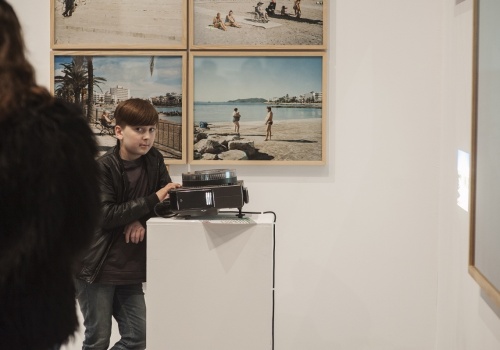 Zdjęcie, zbliżenie na młodego chłopca, który zatrzymał się przy rzutniku. Chłopiec patrzy wprost w obiektyw. Za nim znajdują się prace artysty prezentowane na ścianie.