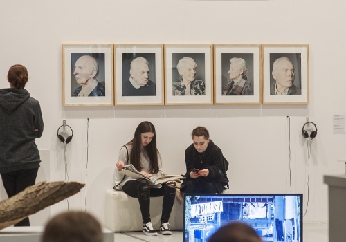 Zdjęcie, pod ścianą, na której prezentowane są prace Michała Szlagi, siedzą dwie uczestniczki wernisażu. Jedna z nich przegląda katalog. Wokół znajdują się inni uczestnicy.