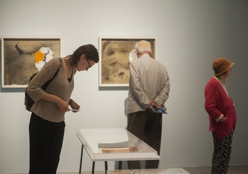Zdjęcie, kilkoro uczestników wernisażu ogląda prace artystki prezentowane w gablotach i na ścianach Sali Wystawienniczej.