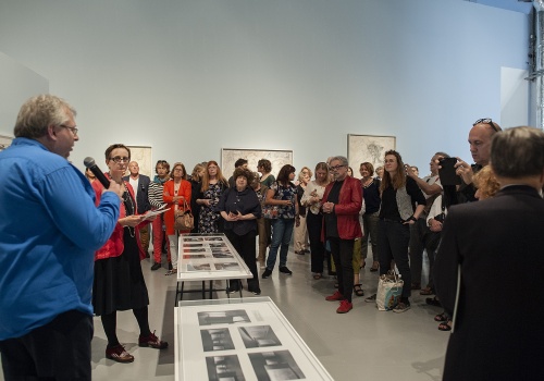 Zdjęcie, licznie zabrani uczestnicy wernisażu w Sali Wystawienniczej, wsłuchują się w wypowiedź mężczyzny, znajdującego się po lewej stronie kadru. Na środku kadru znajdują się szklane gabloty z pracami artystki.