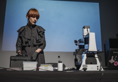 4.	Kobieta w czarnym płaszczu i czarnych rękawiczkach rozstawia mikroskop i inną aparaturę laboratoryjną na ciemnej sali wystawienniczej. 