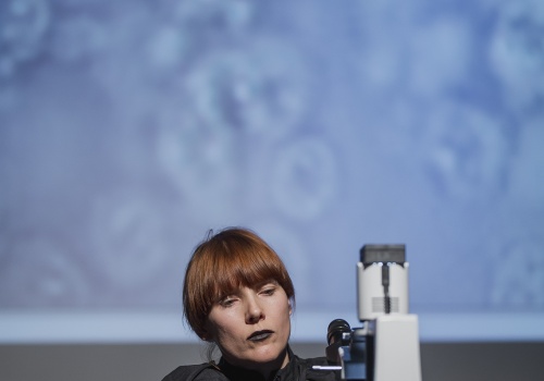 10.	Kobieta w czarnym płaszczu i w czarnych rękawiczkach, znajdująca się na ciemnej sali wystawienniczej, nastraja mikroskop. 