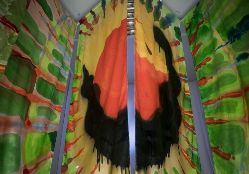 Fotografia pozioma przedstawiająca fragment pracy „Wonsz” wewnątrz kontenera LKW Gallery. Na zdjęciu widać pionowe panele z tkaniny, pomalowane na jasne, żywe kolory. W centralnej części obrazu czarny wąż oplatający czerwone jabłko, a wokół pomarańczowe i zielone promienie na jasnym tle.