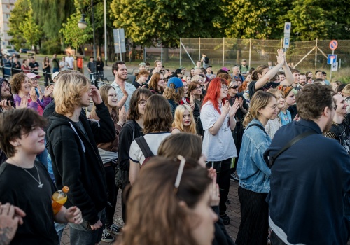 Fotografia pozioma pokazująca tłum uczestników koncertu – klaszczących i radosnych. Są to młodzi ludzie. Niektóry mają kolorowe włosy. Niektóry wznoszą ręce do góry. Jest letni słoneczny dzień. W dalszym planie widać szpaler zielonych drzew i ulicę. 