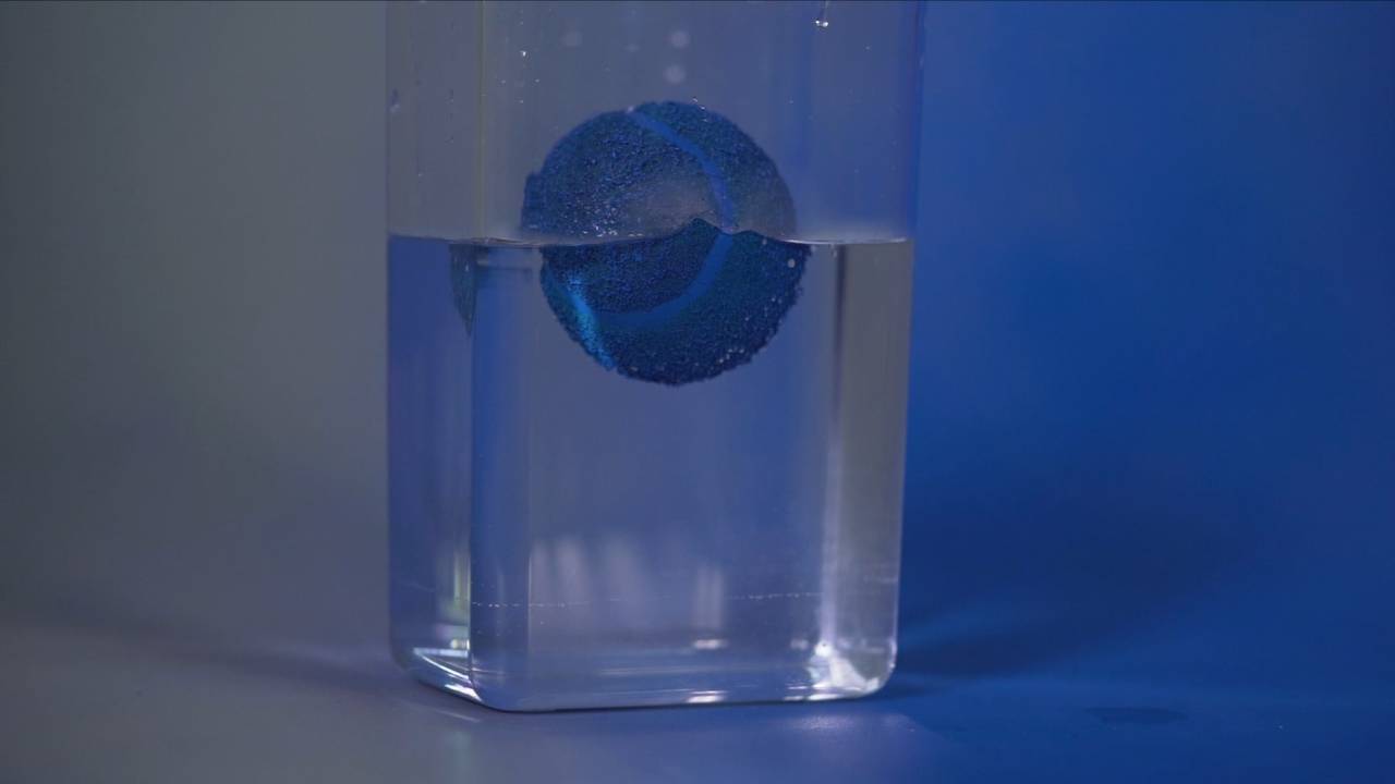 Fotografia pozioma, z centralnie umiejscowionym wysokim, szklanym pojemnikiem do połowy wypelnionym przeźroczystym płynem, w którym zanurzona jest zabarwiona na niebiesko piłeczka do tenisa. Lewa strona fotografii jest zachowana w szarym kolorze, który gradientowo przechodzi w granat, zbliżając się do krawędzi lewej.