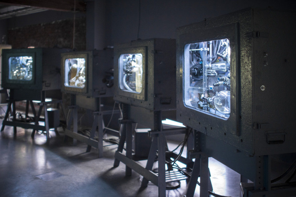 CZtery inkubatory przypominające stare telewizory kineskopowe w metalowej obudowie. Oświetlone wnętrze inkubatorów z licznymi rurkami i przewodami. Wszystkie są ustawione w industrialnej przestrzeni.
