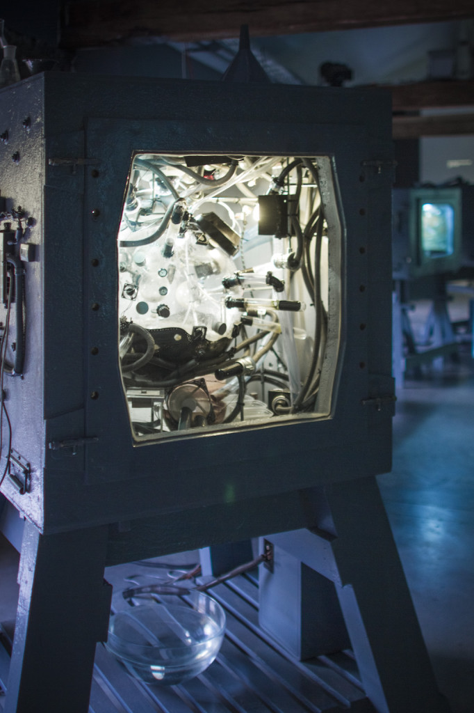 Zbliżenie na inkubator przypominający stary telewizor kineskopowy w metalowej obudowie. Oświetlone wnętrze inkubatora z licznymi rurkami i przewodami. 