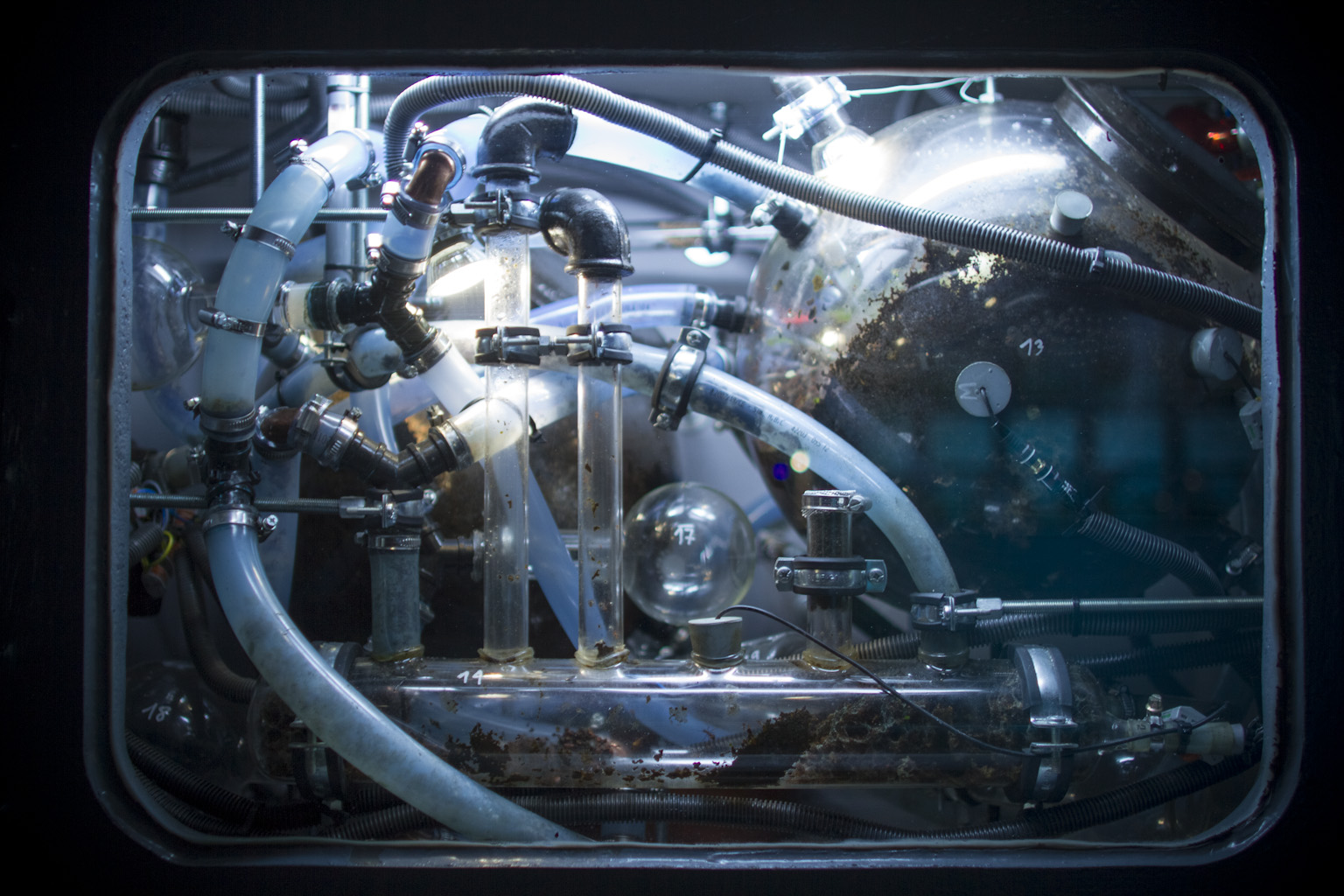 Zbliżenie na inkubator przypominający stary telewizor kineskopowy w metalowej obudowie. Oświetlone wnętrze inkubatora z licznymi rurkami, kolbami i przewodami. 
