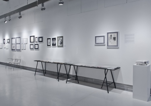 Zdjęcie, zbliżenie na jedną ze ścian Sali wystawienniczej, na której prezentowane są prace artysty. Pod ścianą ustawione są dwie szklane gabloty.