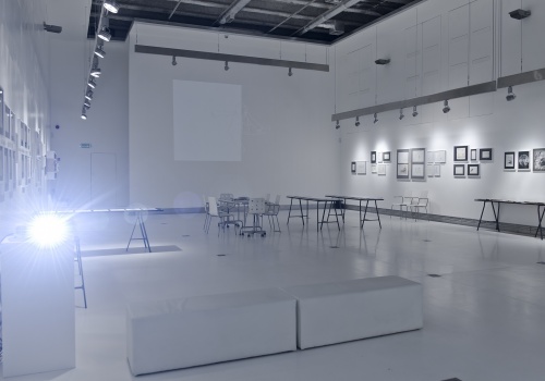 Zdjęcie, wnętrze Sali wystawienniczej. Na ścianach oraz szklanych gablotach, które porozstawiane są na obrzeżach Sali, znajdują się prace artysty. Po lewej stronie kadru znajduje się włączony projektor, który rzuca intensywnie jasne światło.