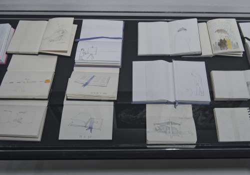 Zdjęcie, zbliżenie na szklaną gablotę. W środku znajdują się pojedyncze kartki lub notatniki z ilustracjami artysty.