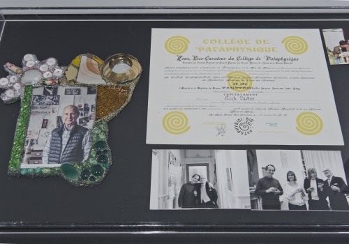 Zdjęcie, zbliżenie na szklaną gablotę. W środku znajdują oficjalne lub prywatne się zdjęcia artysty oraz pamiątkowy dyplom z Patafizyki.