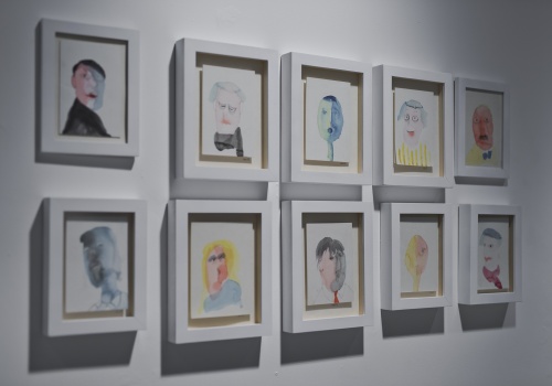 Zdjęcie, zbliżenie na kilka ilustracji artysty, które prezentowane są na ścianie Sali wystawienniczej. Ilustracje przedstawiają kolorowe portrety różnych postaci o różnej mimice.