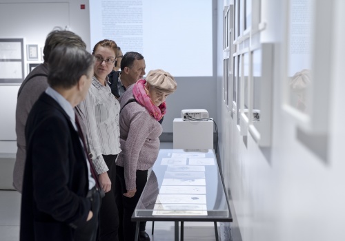 Zdjęcie, zbliżenie na uczestników wernisażu. Jedna z kobiet ogląda prace prezentowane w szklanej gablocie. Pozostali uczestnicy oglądają ilustracje wiszące na ścianie Sali wystawienniczej lub rozmawiają ze sobą.