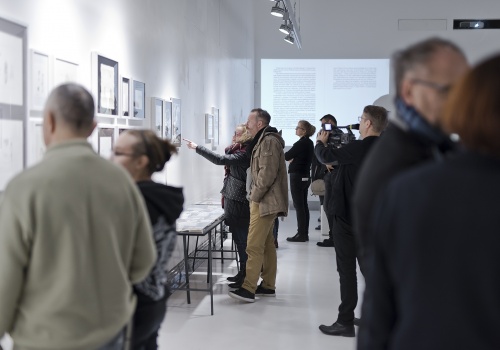 Zdjęcie, wnętrze Sali wystawienniczej podczas trwania wernisażu. Licznie zebrani uczestnicy oglądają prace artysty prezentowane na ścianach oraz w szklanych gablotach. 