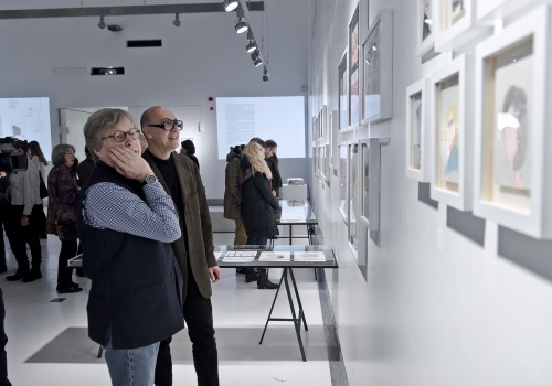 Zdjęcie, zbliżenie na dwójkę uczestników wernisażu, którzy oglądają prace prezentowane na ścianie Sali wystawienniczej. W tle znajdują się pozostali uczestnicy wydarzenia.