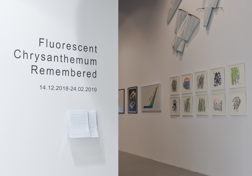 Zdjęcie, dokumentacja wernisażu wystawy japońskich artystów pod tytułem „Fluorescent Chrysanthemum Remembered”. Wejście na Salę Wystawienniczą CSW ŁAŹNIA 1 na Dolnym Mieście. Po lewej stronie kadru znajduje się biała ściana, a na niej czarne napisy – tytuł wystawy oraz data 14.12.2018-24.02.2019. Po prawej stronie znajduje się wnętrze Sali Wystawienniczej wraz z zaprezentowanymi pracami artystów.