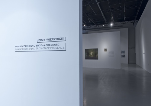 Zdjęcie, dokumentacja wernisażu wystawy Jerzego Wierzbickiego pod tytułem „Oman/Czarnobyl. Erozja obecności”. Wejście na Salę Wystawienniczą CSW ŁAŹNIA 1 na Dolnym Mieście. Po lewej stronie kadru znajduje się biała ściana, a na niej czarne napisy – imię i nazwisko artysty oraz tytuł wystawy w języku polskim i angielskim. Po prawej stronie znajduje się wnętrze Sali Wystawienniczej wraz z zaprezentowanymi pracami artysty.