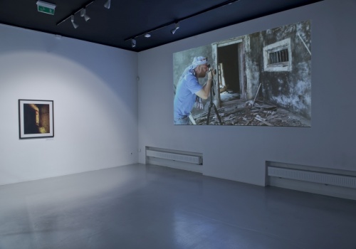 Zdjęcie, wnętrze  Sali Wystawienniczej. Na ścianie po lewej stronie prezentowana jest fotografia Jerzego Wierzbickiego, a po prawej stronie wyświetlany jest film artysty. W zatrzymanym kadrze znajduje się Wierzbicki fotografujący fragment budynku.