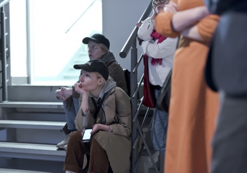Zdjęcie, dwie uczestniczki wernisażu siedzą na schodach prowadzących do wnętrza Sali Wystawienniczej. Oglądają film artysty, który wyświetlany jest przed nimi na ścianie, która znajduje się poza kadrem.