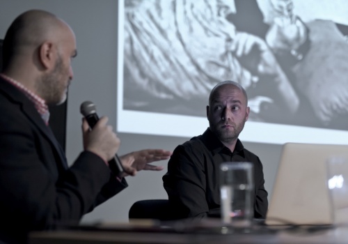 Zdjęcie, wnętrze Parakina. W centrum kadru znajduje się Jerzy Wierzbicki, który słucha wypowiedzi mężczyzny, znajdującego się po lewej stronie. Mężczyzna ten mówi do mikrofonu. W tle znajduje się ściana, a na niej wyświetlana jest czarno biała fotografia.