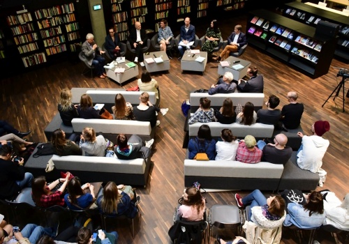 Zdjęcie, widok z góry na osoby prowadzące dyskusje. Przed nimi znajdują się zebrani słuchacze. Za nimi widnieje długi, ciemny regał z książkami.