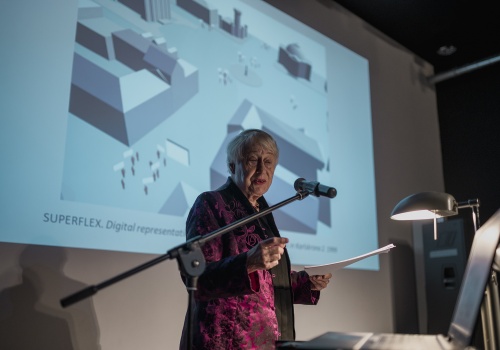 Zdjęcie, konferencja pod tytułem „Miasto i Sztuka”. Jedna  z zaproszonych kobiet wypowiada się do mikrofonu. Za nią wyświetlana jest prezentacja ze schematem budynków.