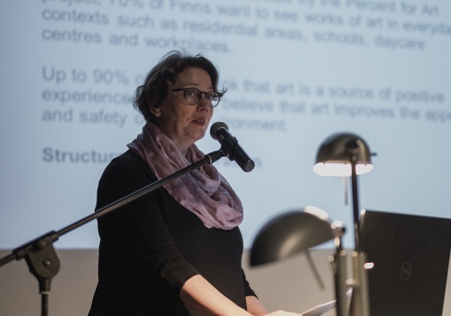 Zdjęcie, konferencja pod tytułem „Miasto i Sztuka”. Jedna z zaproszonych kobiet wypowiada się do mikrofonu. Za nią wyświetlana jest prezentacja z czarnymi napisami w języku angielskim.