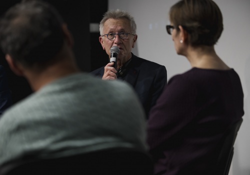 Zdjęcie, zbliżenie na starszego mężczyznę w okularach, wypowiadającego się do mikrofonu. Przed nim znajdują się wie osoby, siedzące tyłem do widza, wśród nich Dyrektor CSW ŁAŹNIA – Jadwiga Charzyńska.