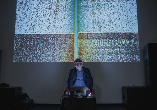 4.	Mężczyzna w wieku średnim siedzi przed laptopem ustawionym na niewielkim stoliku. Na ścianie za mężczyzną wyświetlana jest wielkoformatowa praca wizualna.Zdjęcie, jeden z artystów, znajdujący się w centrum kadru, prezentuje swoją pracę – wizualizację, która wyświetla się na ścianie, za jego plecami, w ciemnej Sali Parakina. 