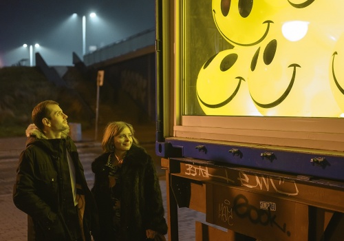 Dwie postacie oglądające widoczną w prawej części fotografii instalację „Smile, please”, skonstruowaną z podświetlonych, żółtych balonów z nadrukiem uśmiechniętego emotikonu. Instalację  autorstwa Magdaleny i Ksawerego Kirklewskich umieszczono we wnętrzu galerii LKW